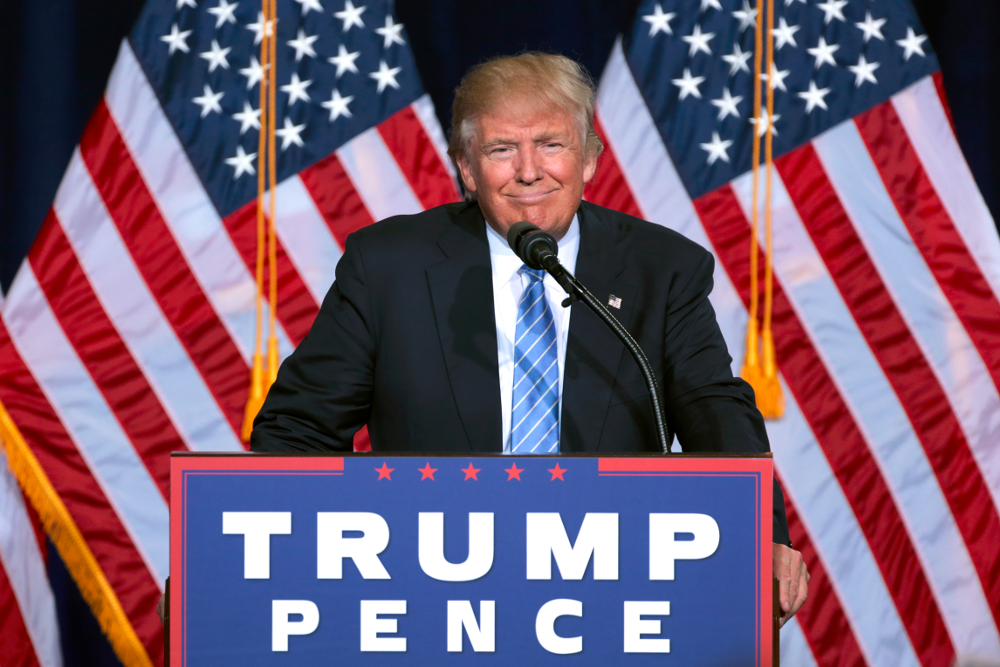 Trump standing behind a brown wood podium with a sign bearing the words "TRUMP PENCE"