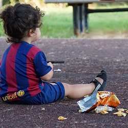 child in red and blue striped shirt sitting on the ground eating junk food potato chips
