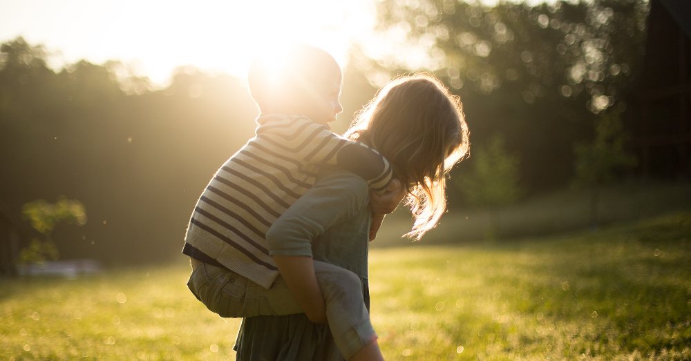 two children playing piggyback in a grassy field at sunset