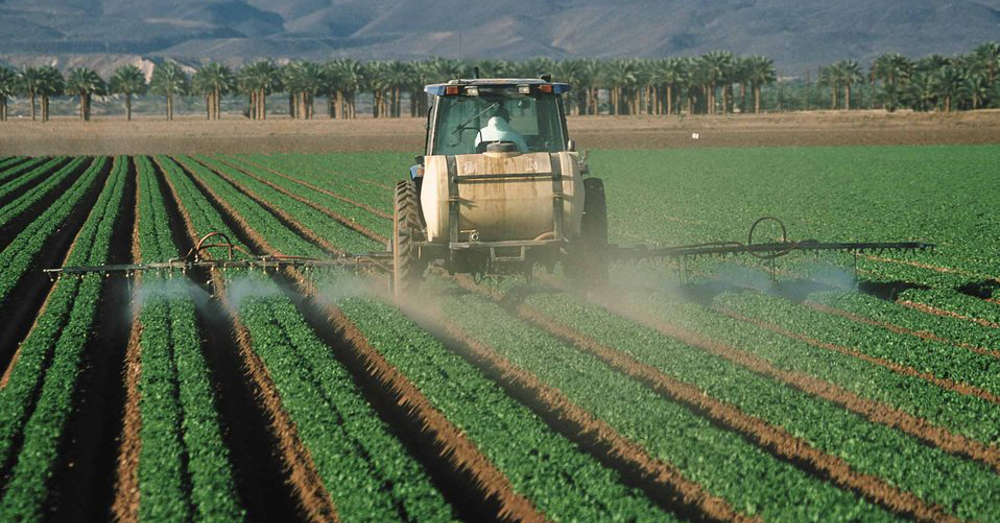farmer in a tractor applying a spray herbicide or pesticide to a row of agricultural crops