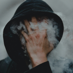person holding their hand over their hooded face with smoke circling