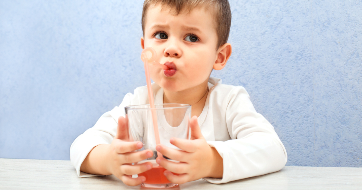 young boy drinking grape juice out of a glass with a straw against a blue background
