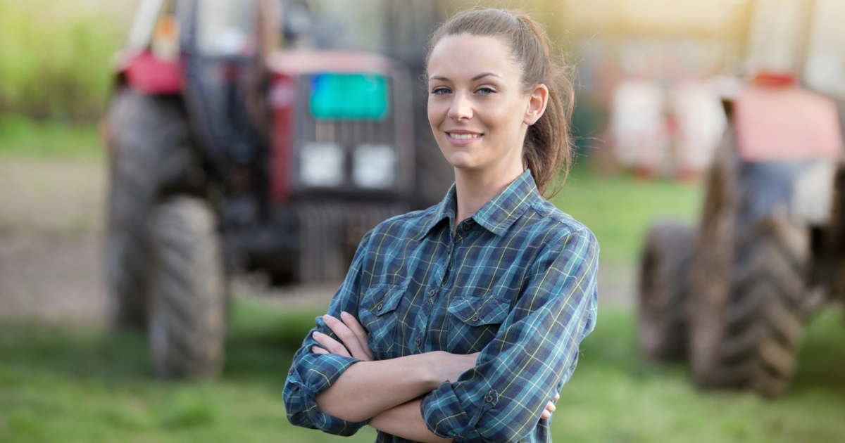 woman rancher stands with arms crossed in front of several tractors on a farm field