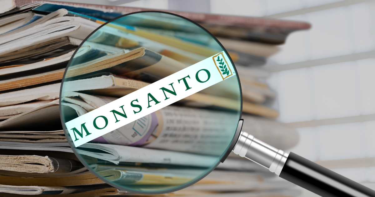 Magnifying glass with Monsanto logo underneath with stack of newspaper in backgroun