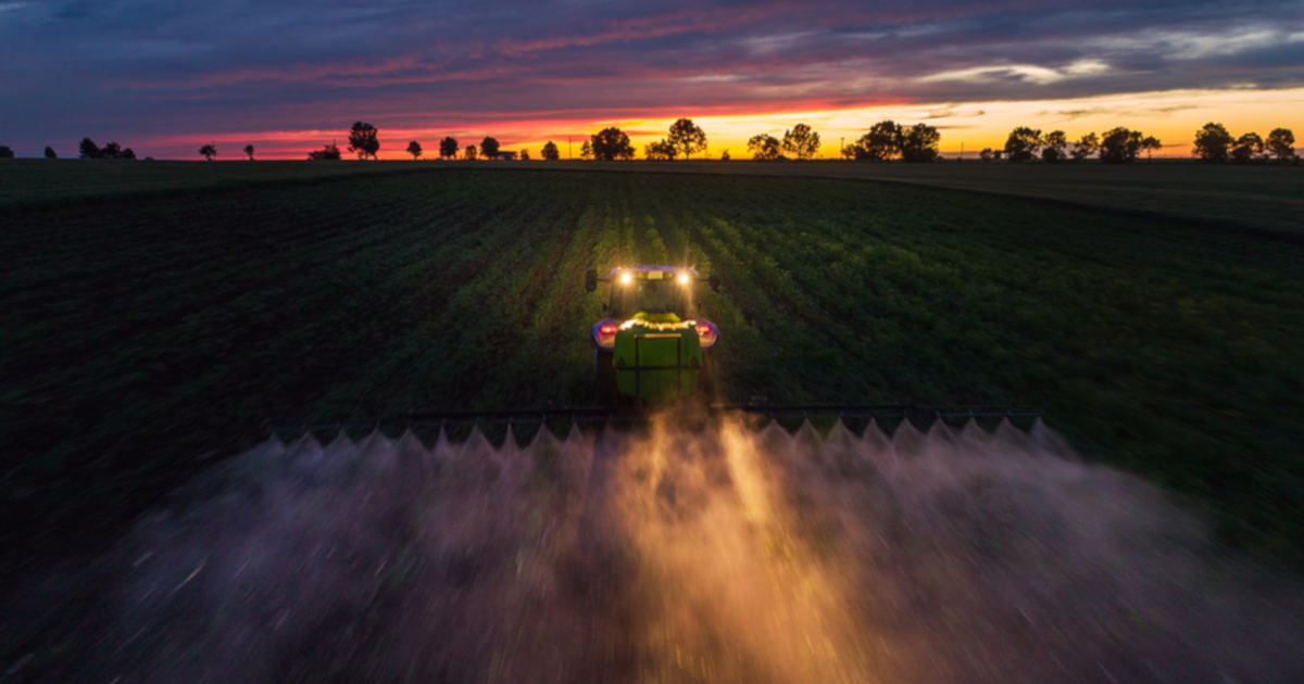 tractor with its lights on spraying herbicide on farm field crops at sunset