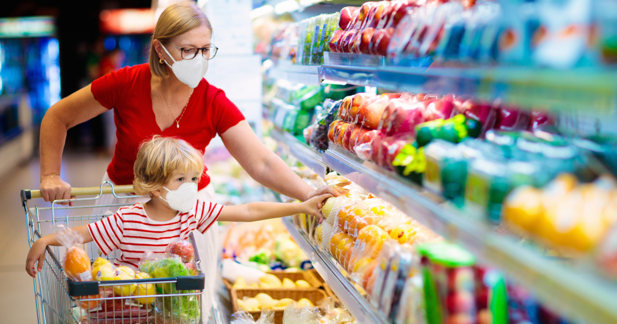 mother and young son shopping in the produce aisle of a supermarket wearing masks