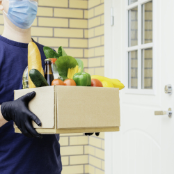 man in a mask and gloves delivering a box of vegetables and other produce to a persons door