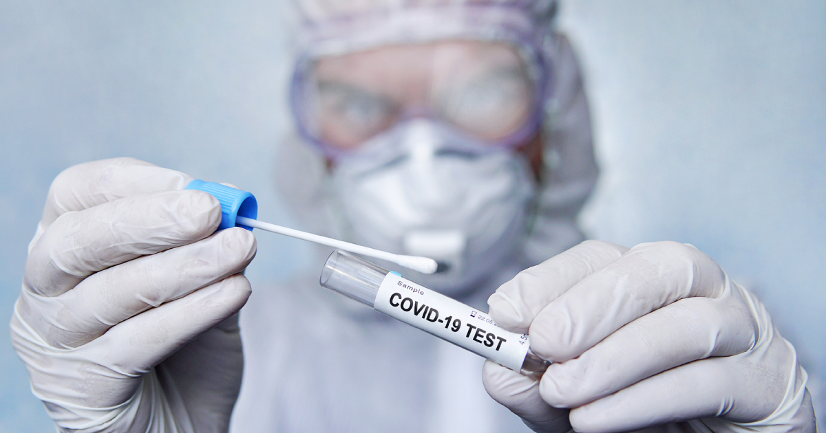 A photograph of a scientist wearing a hazmat suit, goggles and gloves holding a test tube that says "COVID-19 test"